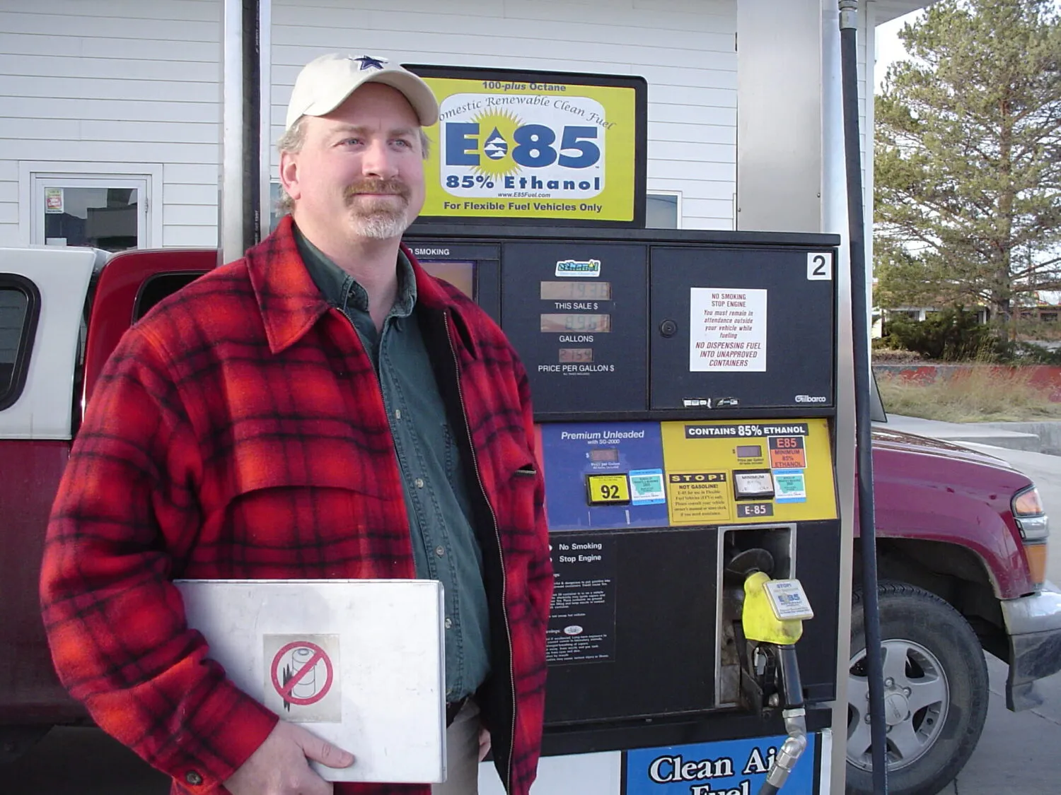 Man in a red plaid jacket standing next to a gas pump, holding a metal container.
